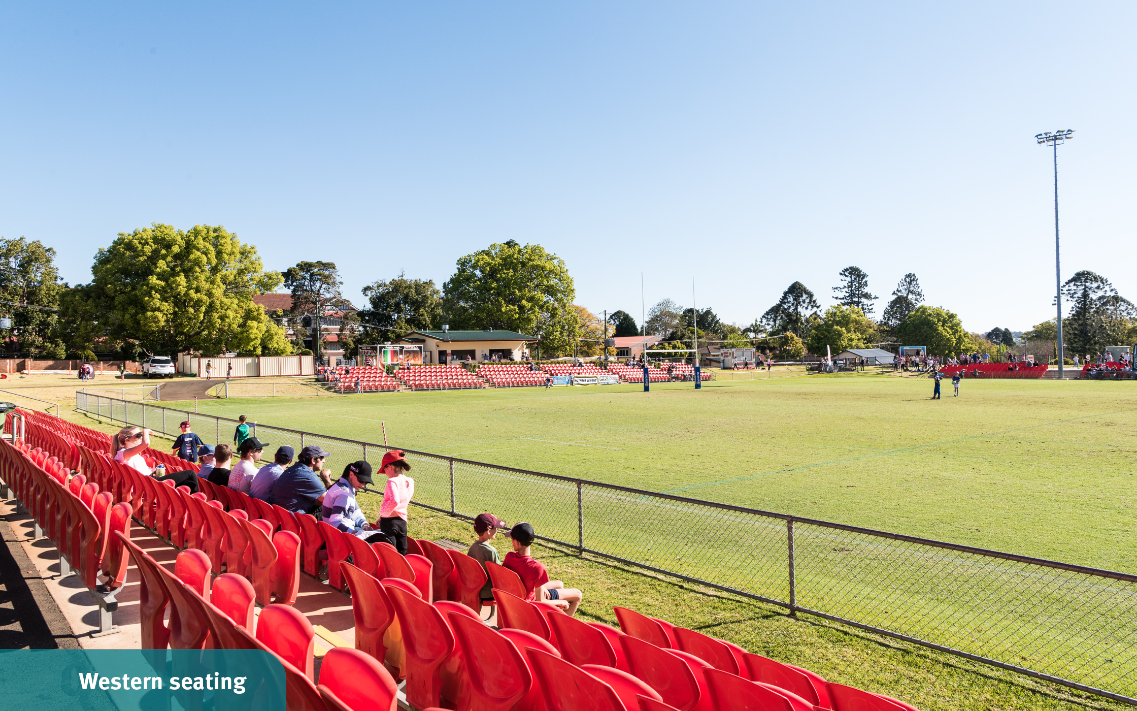 Spectators sitting in red outdoor seating watching the green field as they wait for a football game to begin at the Toowoomba Sports Grounds