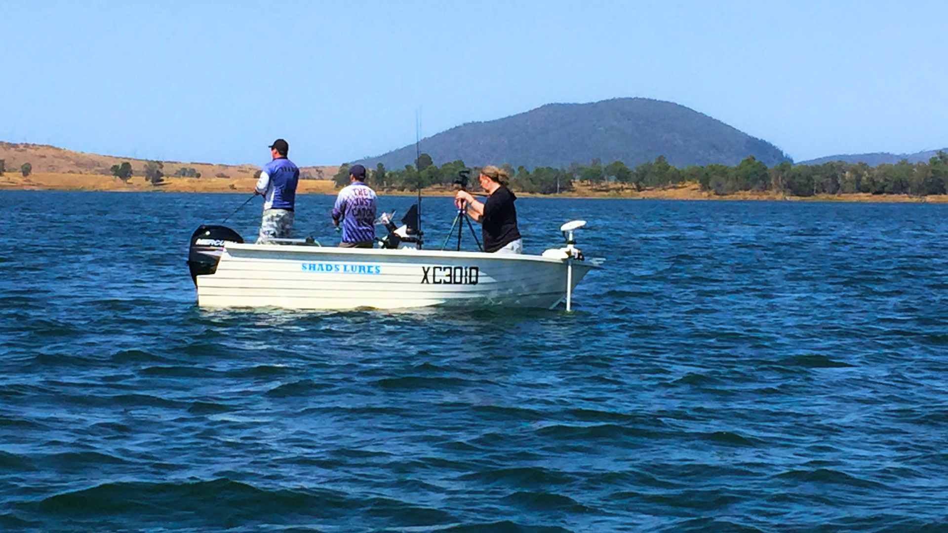 Anglers fishing from a small boat on open blue water with hills behind.