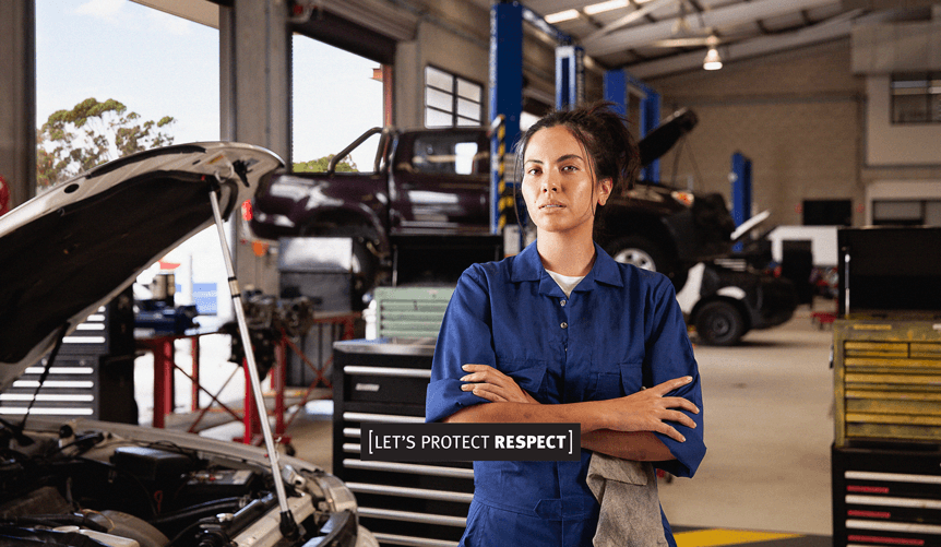 girl in overalls stands with arms folded looking serious in a mechanics workshop