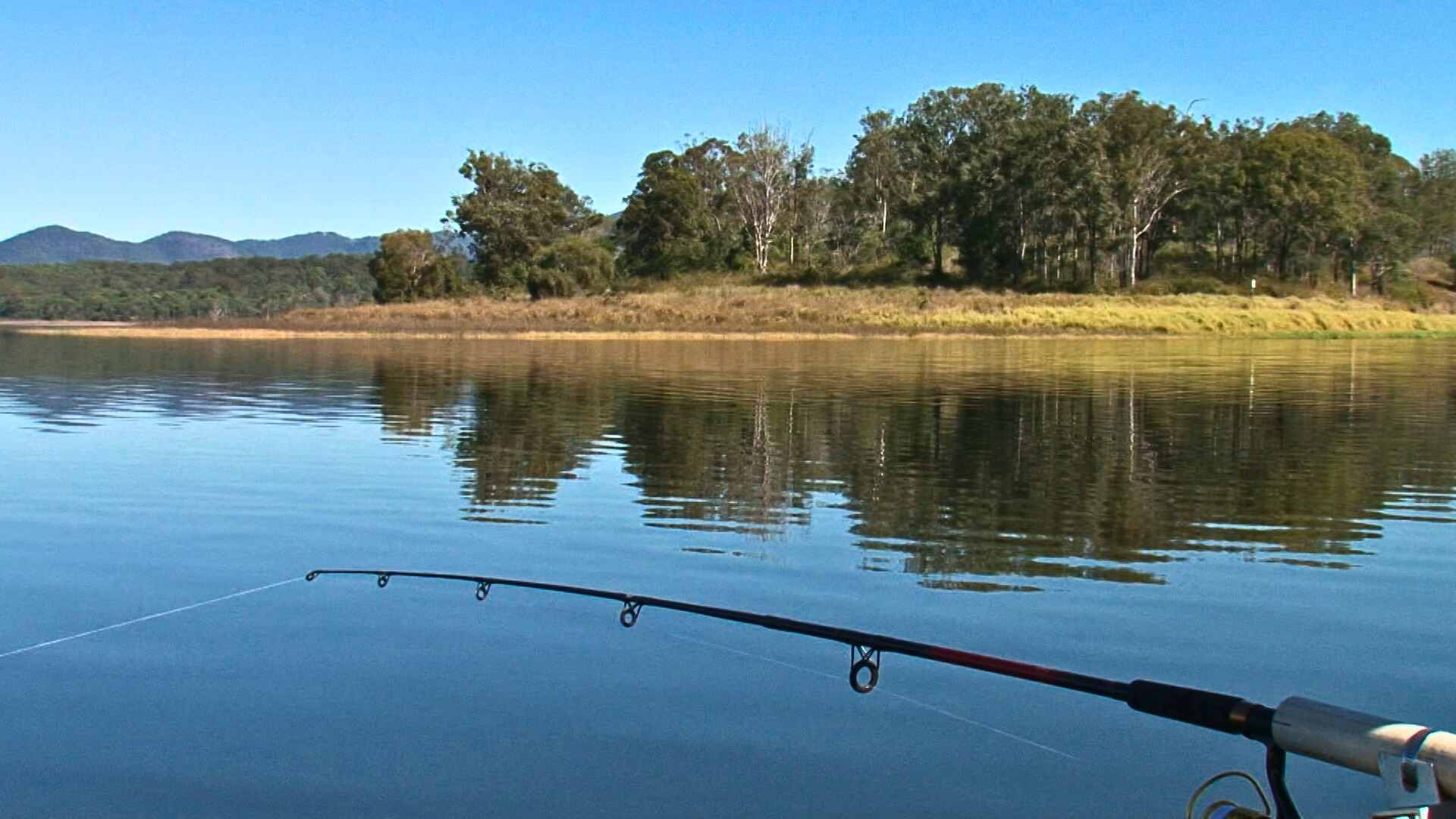 Fishing rod resting over still water reflecting trees and hills.