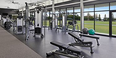 Interior view of the gym showing weight machines, benches, and a view through full sized windowsto a green field.