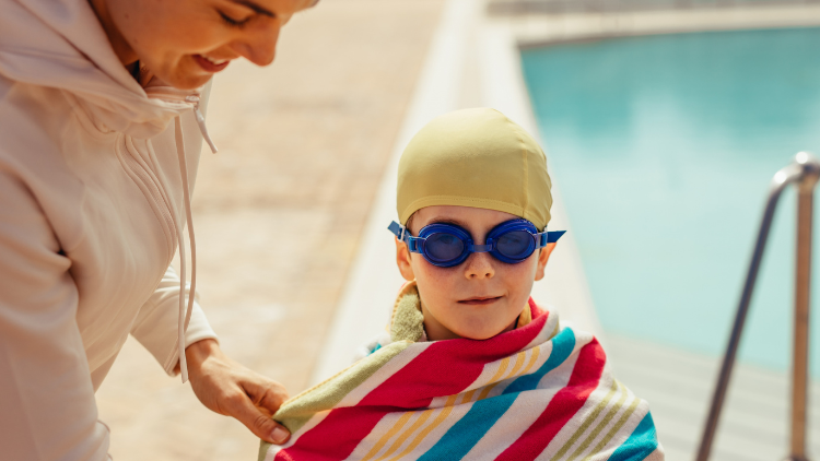Child with swimming cap and googles standing by the pool in a towel with an adult