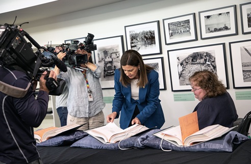 Minister Deb Frecklington stands over a table with large documents that have been opened. A woman sits to her side and men with video cameras are also standing around the table.