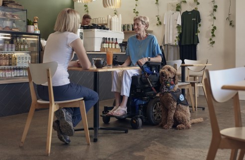 A woman in a wheelchair is accompanied by an assistance dog. She is in a café with a friend.