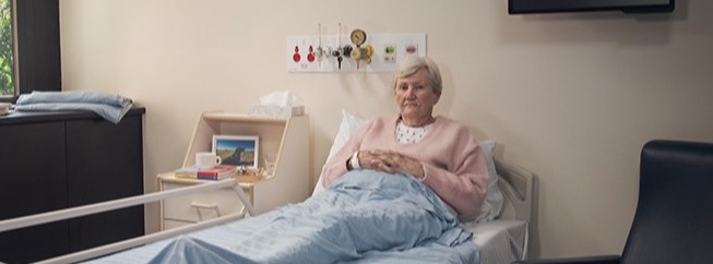 An elderly woman sits upright in a hospital bed in a private room, covered with a light blue blanket. The room has a window with trees outside, a wall-mounted TV, a bedside tray with a cup and framed photo, and an empty chair beside the bed.