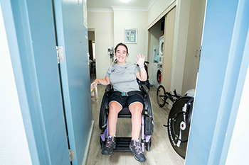 A woman in a wheelchair waves and smiles at the camera